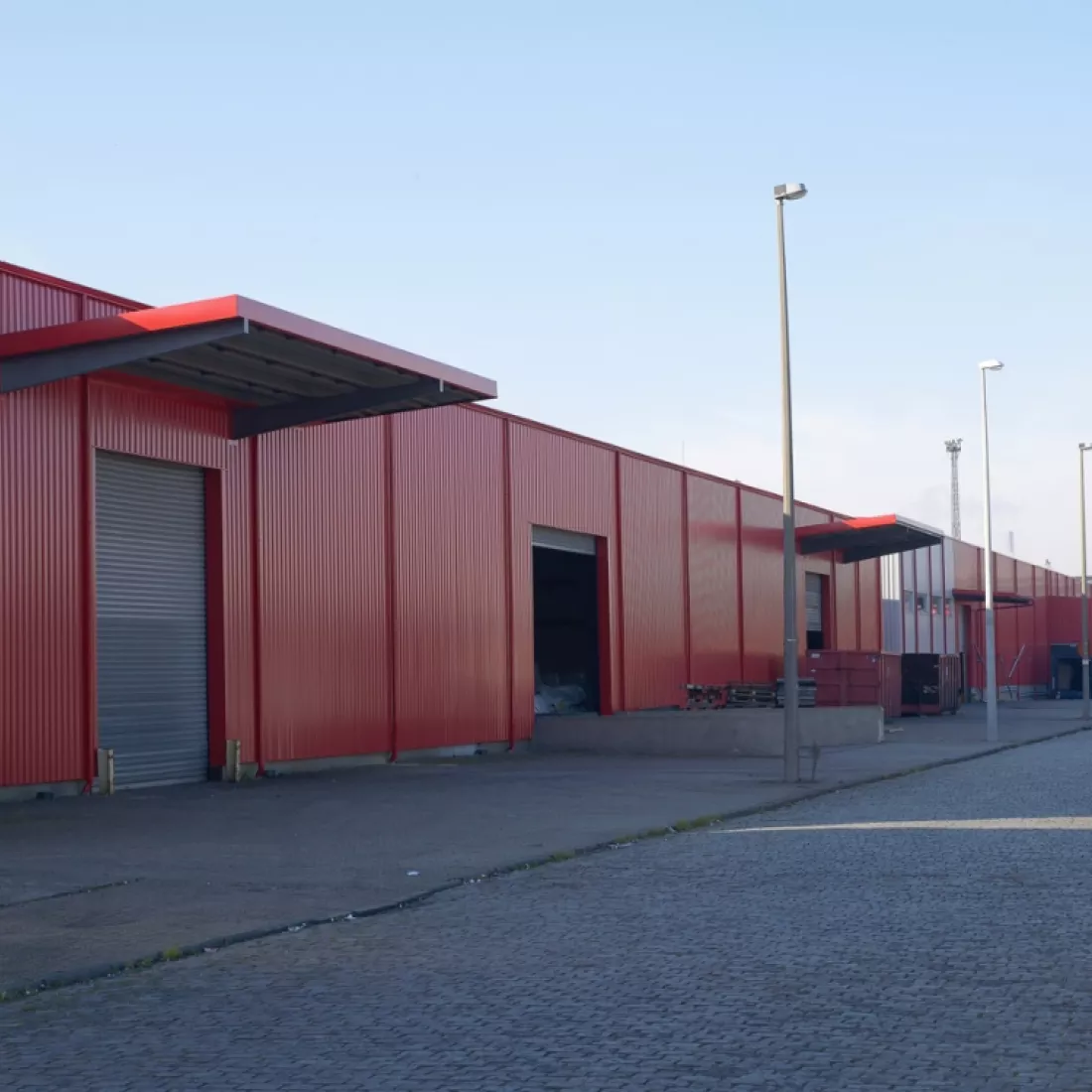 Zijaanzicht renovatie daken en wanden van magazijn TCL. De wanden zijn rood. De poorten zijn grijs. Boven sommige poorten hangt een rode luifel.