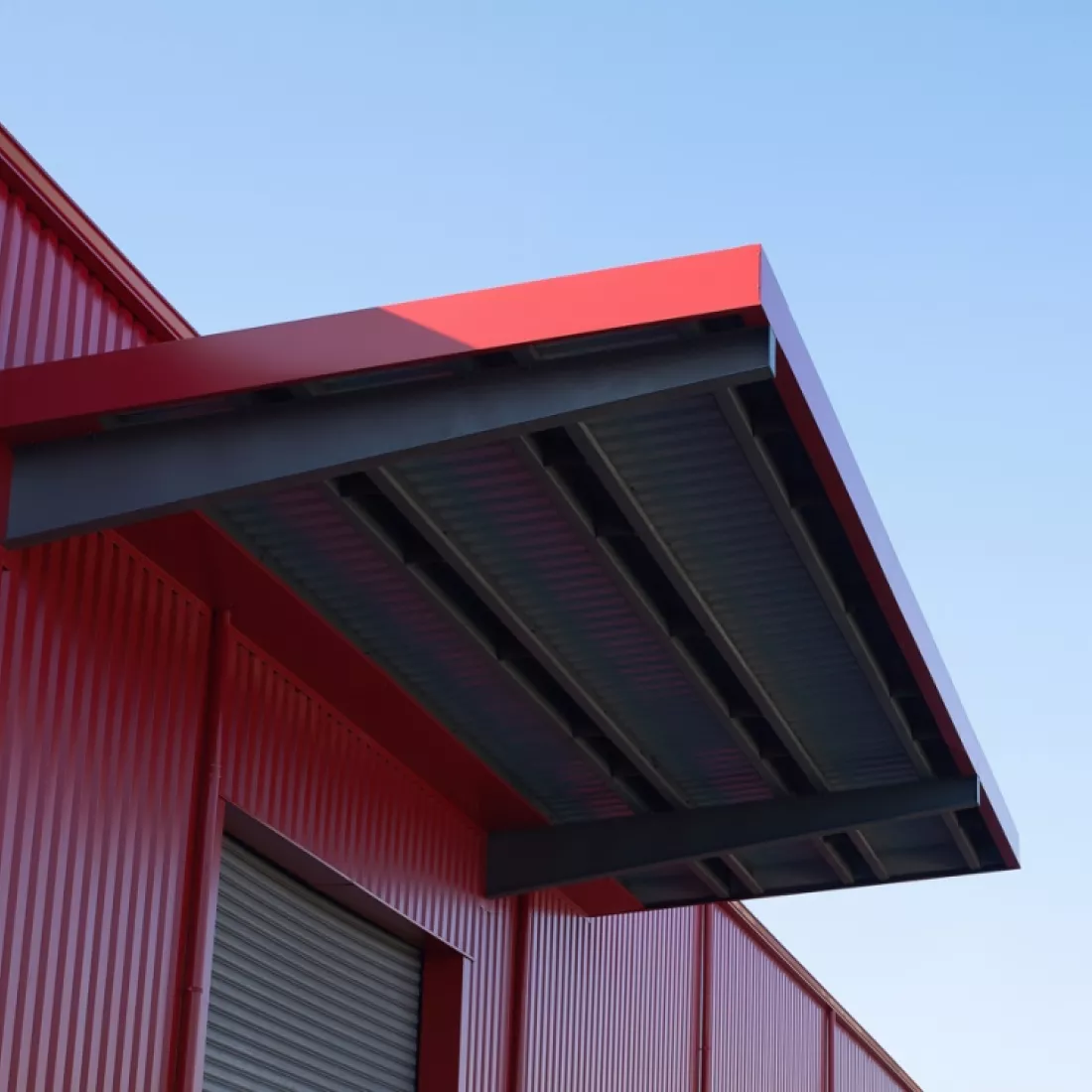 Close-up magazijn TCL. Het is een close-up van een luifel boven de poort. De poort is grijs en de luifel en wanden rood.