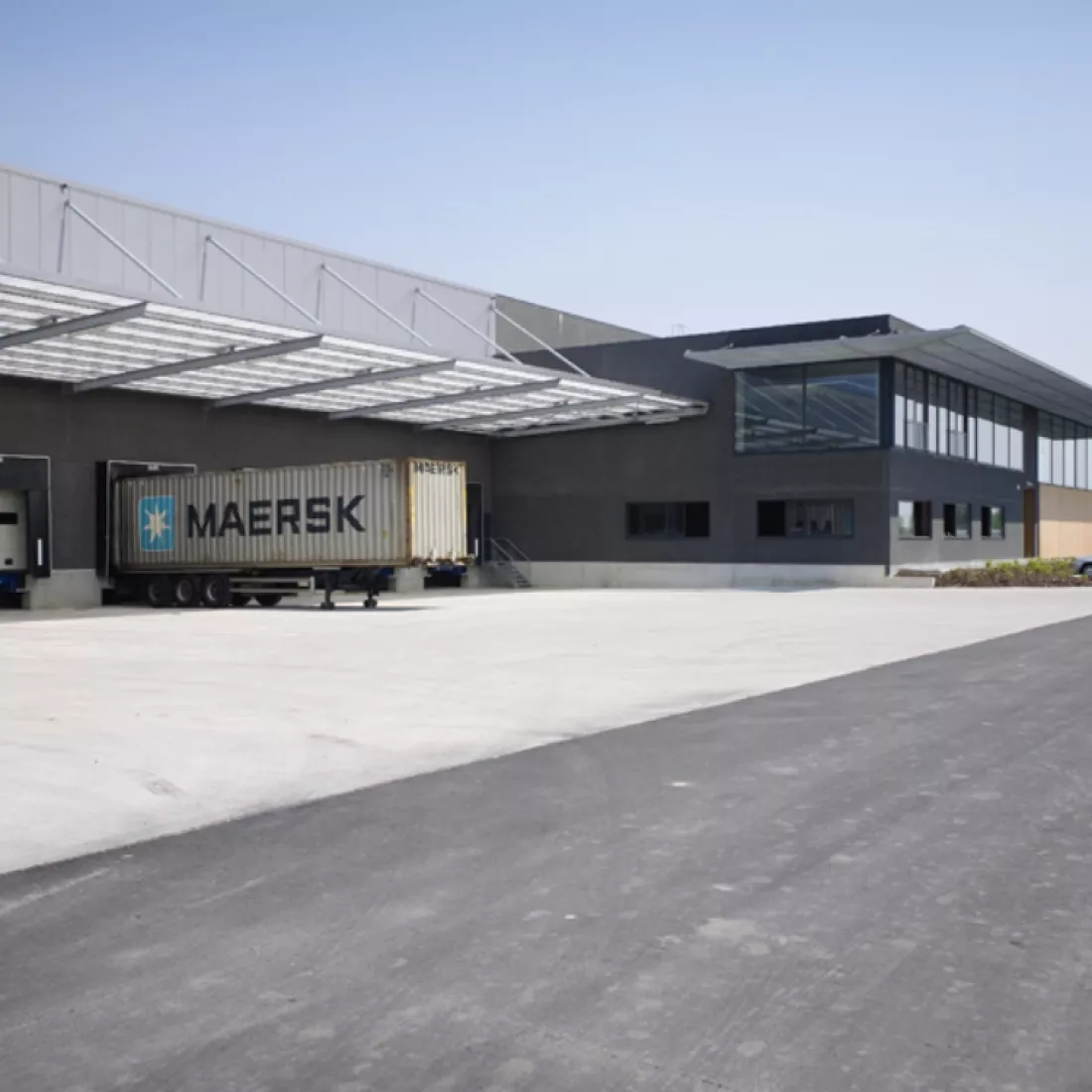 Dock van magazijnruimte met kantoor - Ethnicraft. Hier laden en lossen de vrachtwagens. Het gebouw is opgebouwd met beton. Het dock bestaat uit grijze platen. Rechts naast het dock bestaat het gebouw uit zwarte platen en heeft het gebouw verschillende soorten ramen.