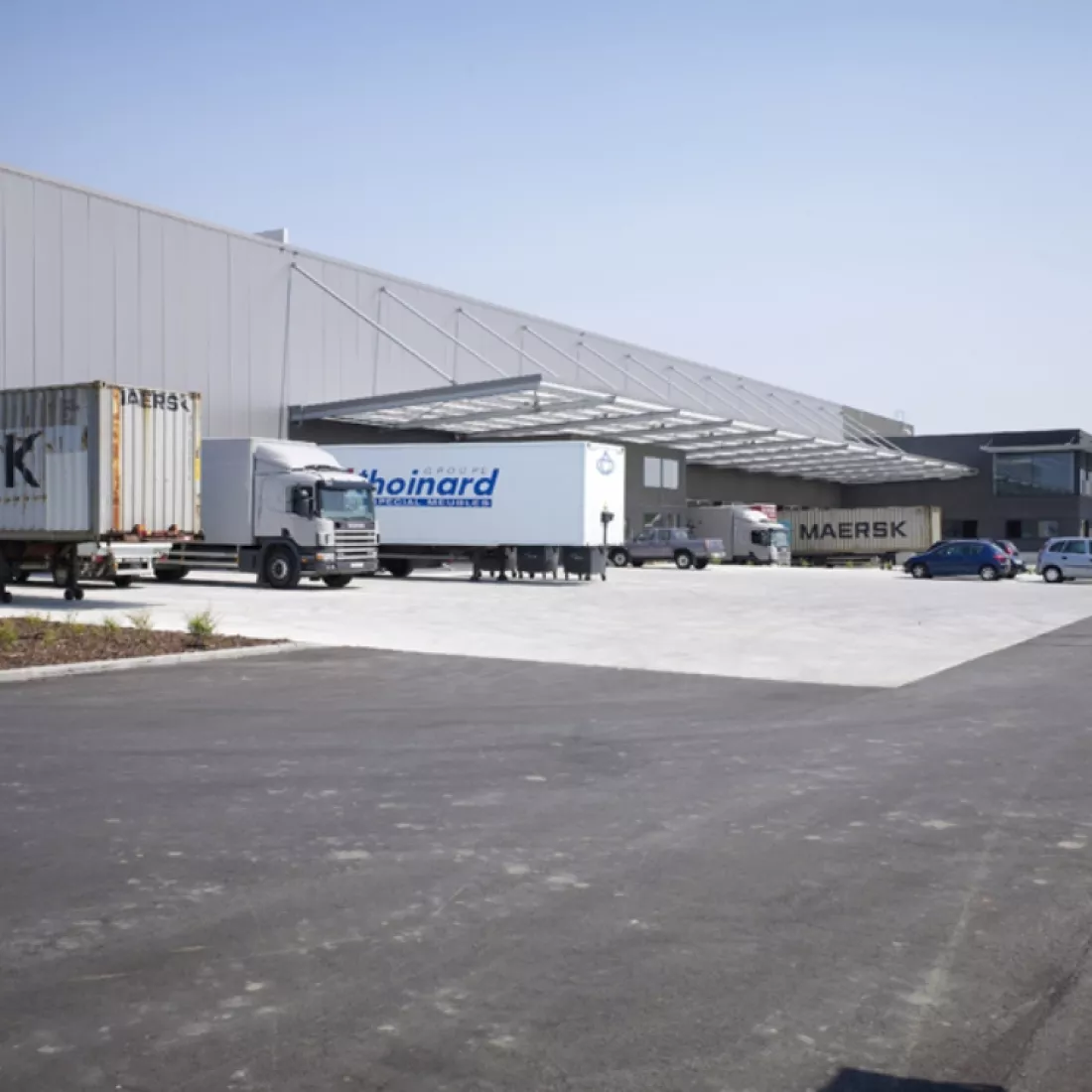 Dock van magazijnruimte met kantoor - Ethnicraft. Hier laden en lossen de vrachtwagens. Het gebouw is opgebouwd met beton. Het dock bestaat uit grijze platen. Rechts naast het dock bestaat het gebouw uit zwarte platen en heeft het gebouw verschillende soorten ramen.