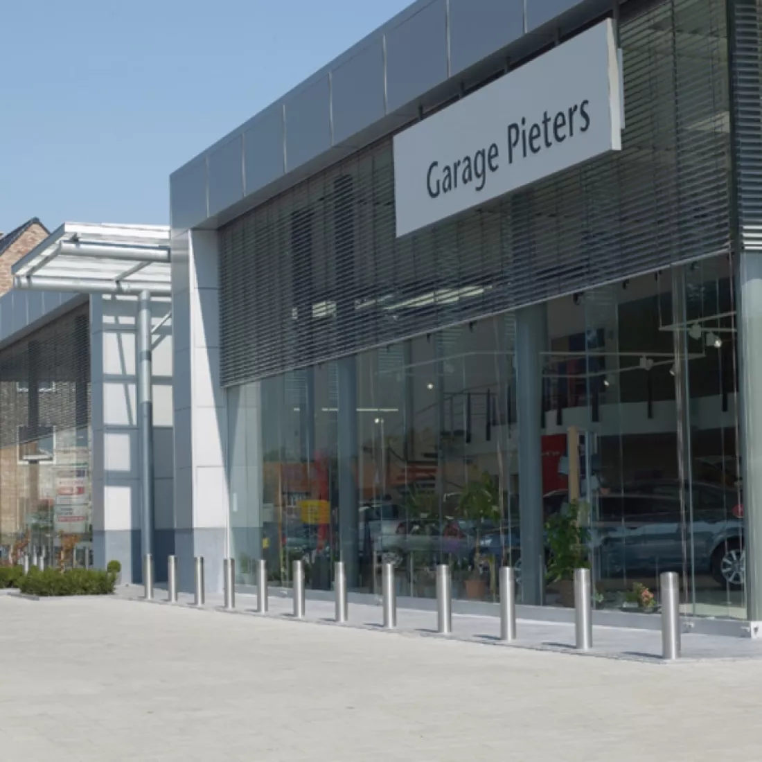 Zijaanzicht showroom garage Pieters. De showroom bestaat uit grote ramen. Tegen het gebouw hangt een wit bord. Op het bord staat 'garage Pieters' in zwarte letters. Voor het gebouw staan stalenpaaltjes. Dit om te voorkomen dat iemand tegen de ramen rijdt.