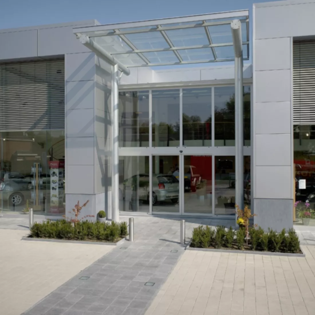 Vooraanzicht inkom showroom garage Pieters. Het gebouw is grijs en bevat veel grote ramen. Voor de inkom hangt een glazen luifel.