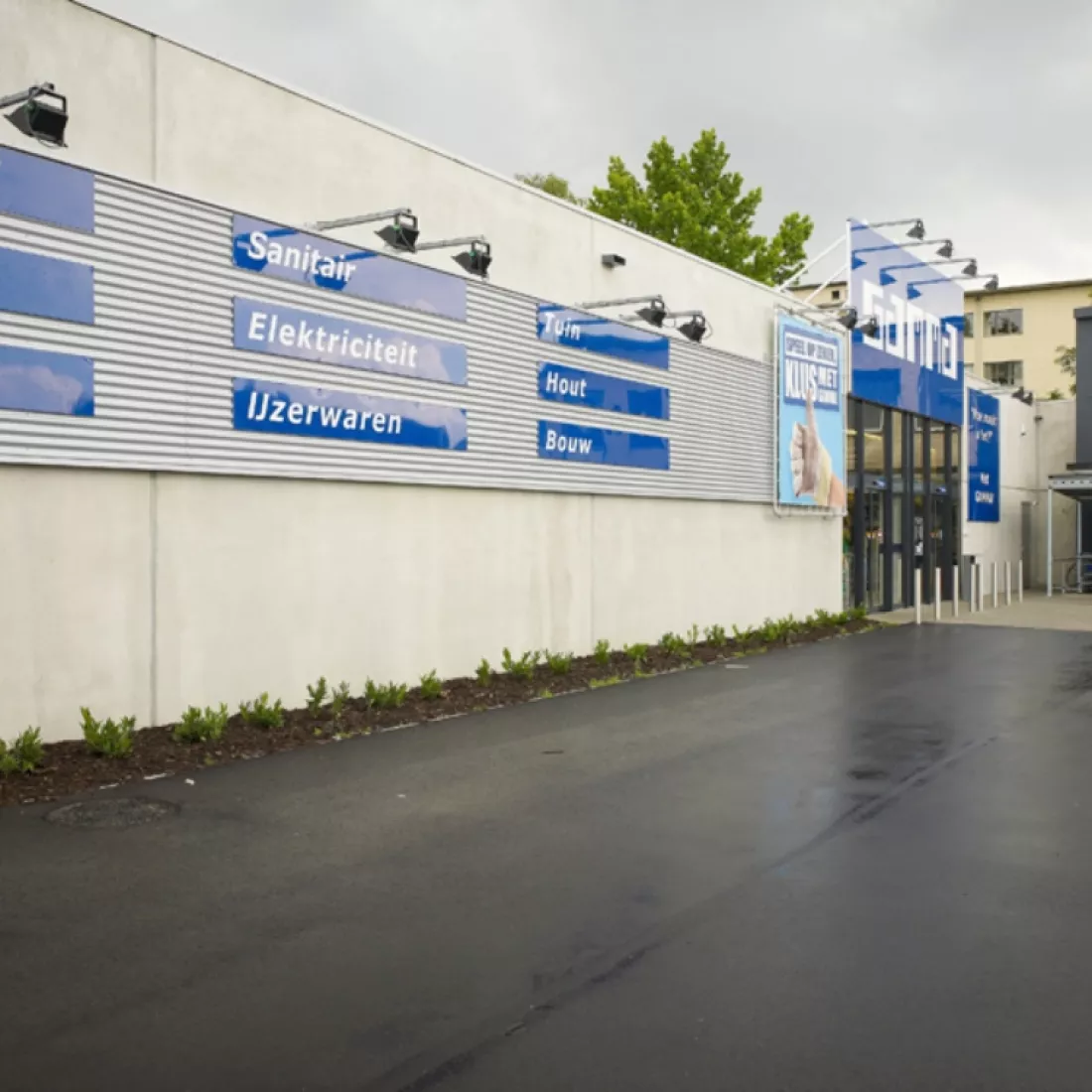 Zijaanzicht winkelruimte Gamma - Trading Places. Het gebouw is grijs. Er zijn blauwe borden die tegen het gebouw hangen. Tijdens de opbouw is er staal gebruikt.