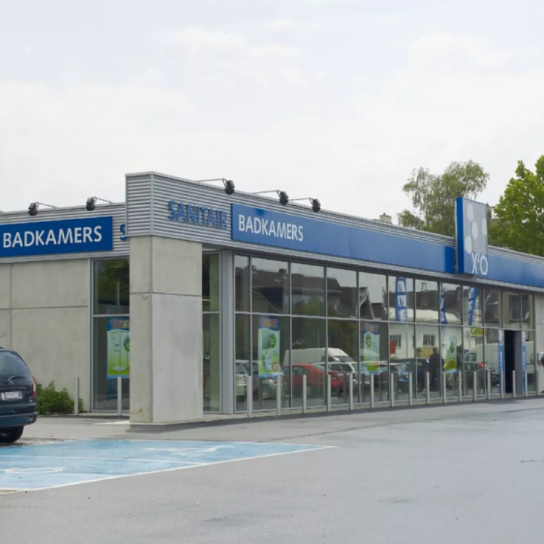 Zijaanzicht winkelruimte X²O - Trading Places. Het gebouw is grijs en heeft aan de voorkant grote ramen. Het eerste deel van de linker muur bestaat ook nog uit ramen. Bovenaan het gebouw hangen blauwe borden en letters van het merk.