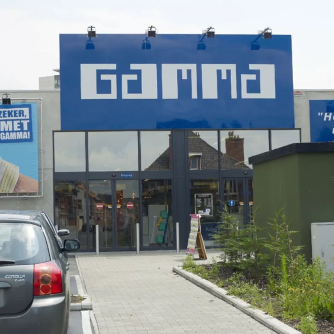 Vooraanzicht winkelruimte Gamma - Trading Places. Het gebouw is grijs. Zowel de in- als uitgang bestaat uit automatische schuifdeuren, deze bestaan uit gals met een zwarte omranding. Boven de deuren hangt een groot bord met het logo van gamma.
