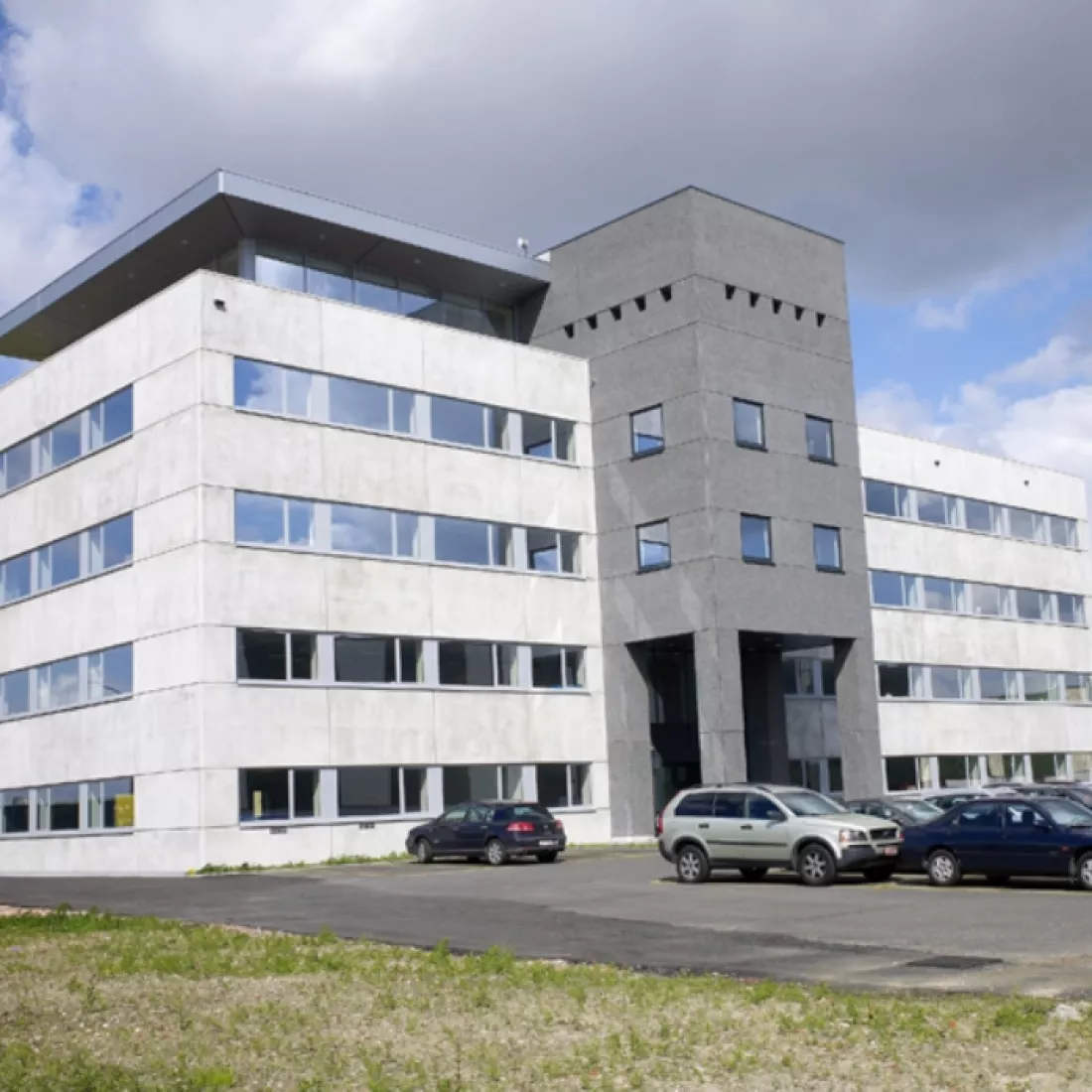 Vooraanzicht kantoorgebouw Nova Natie HQ. Het gebouw is lichtgrijs en in het donkergrijs. Het heeft vijf verdiepingen. Elke verdieping heeft veel ramen. Voor het kantoor is parkeerruimte voorzien.