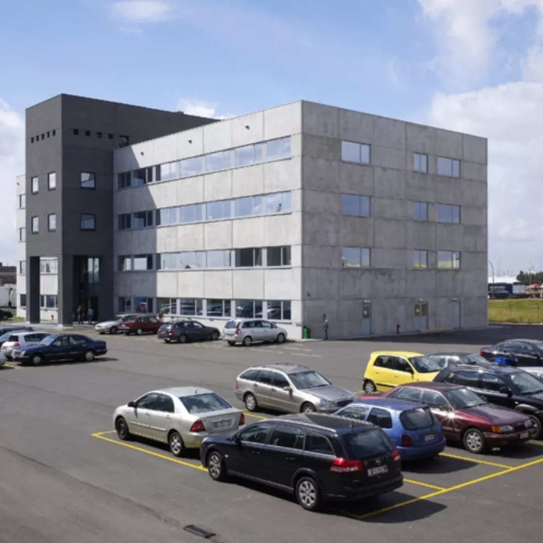 Zijaanzicht kantoorgebouw Nova Natie HQ. Het gebouw is een staalbouw. Voor het gebouw zijn parkeerplaatsen voorzien. Deze zijn afgebakend met gele randen.