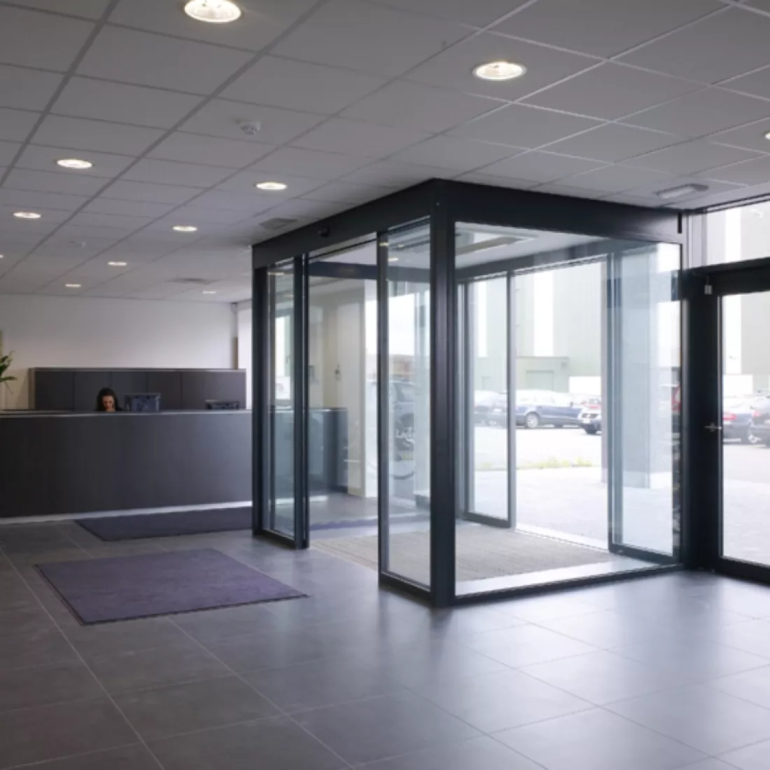 Inkomhal kantoorgebouw Nova Natie HQ. De inkom bevat een dubbeldeursysteem. Rechts van de deur staat een zwarte balie. De muren zijn wit.