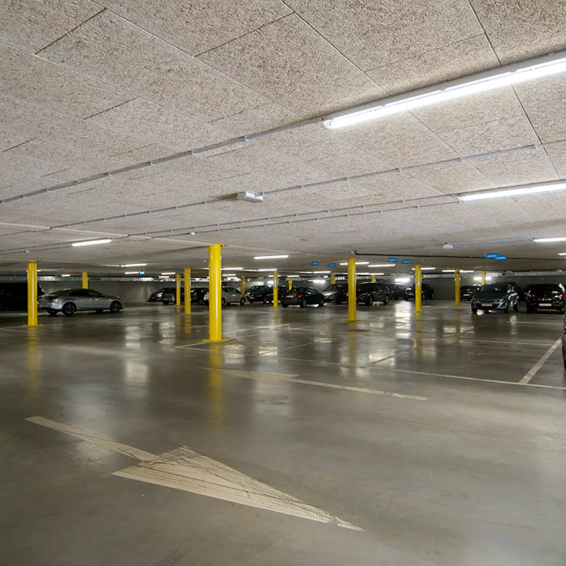 Ondergrondse garage. In de garage zijn parkeerplaatsen afgebakend met witte strepen. Er staan gele pilaren in de garage. Met witte pijlen is de richting aangegeven op de grond. Er staan verschillende auto's geparkeerd. Tijdens de opbouw is er staal gebruikt.