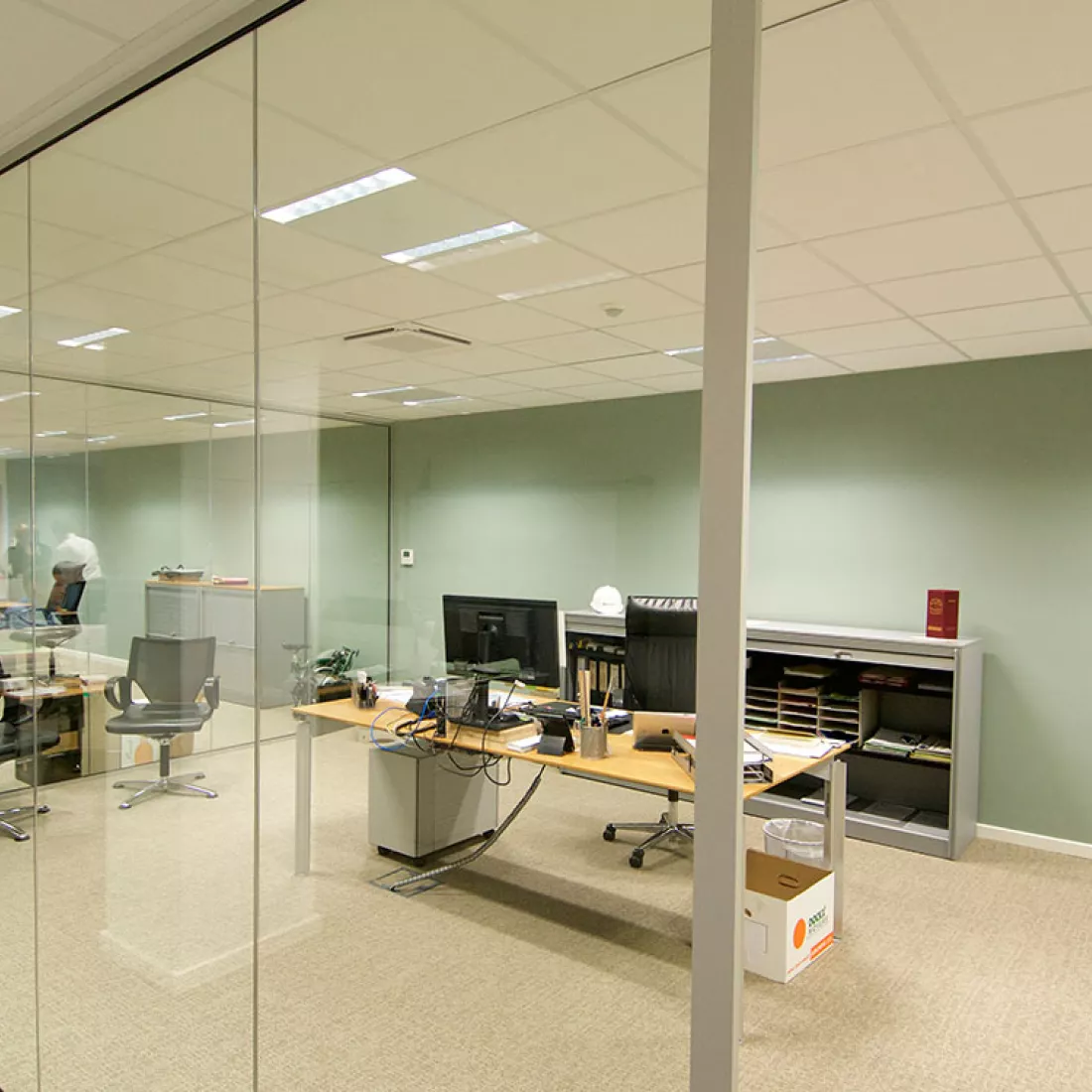 Binnenkant kantoor Altripan. De kantoren zijn van elkaar gescheiden met glazen wanden. De achterste muur heeft een pastelgroene kleur. In het midden staat een bureau. Achter het bureau staat een kast.