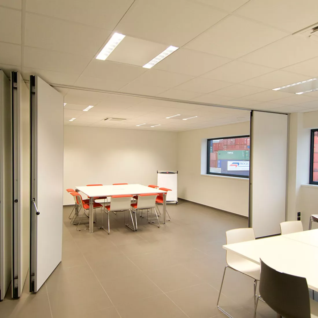 Binnenkant kantoor Altripan. De afbeelding is genomen van een vergaderruimte. De ruimte kan verdeeld worden in twee aparte ruimtes door een witte tussenwand. In de twee delen staat een vierkante tafel met stoelen rond. De rechtermuur bevat ramen.