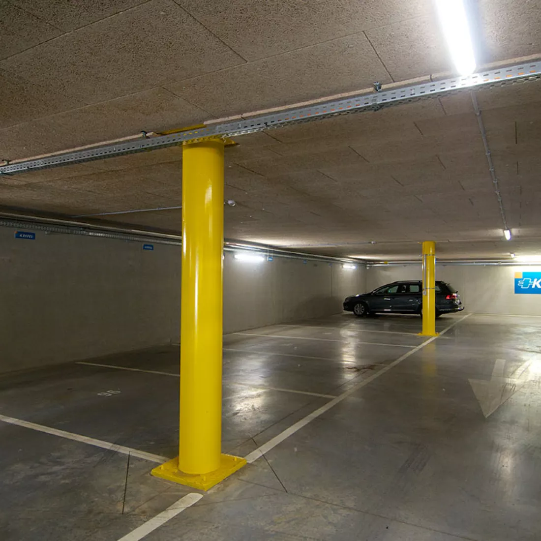 Close-up garage Brussel. Er zijn witte lijnen op de grond voor het afbakenen van parkeerplaatsen. Ook zijn er witte pijlen die de richting aanduiden. Er staan gele pilaren in de garage. Helemaal achteraan staat een auto geparkeerd.