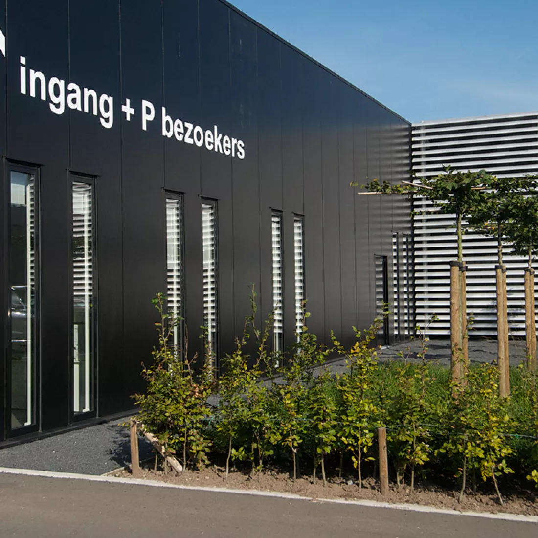 Zijaanzicht toonzaal, kantoren, showroom Vimmo. Het gebouw is zwart met smalle ramen. Tegen het gebouw hangen witte letters die de woorden 'ingang + P bezoekers' vormen. Rechts naast het gebouw is groen zoals bomen.