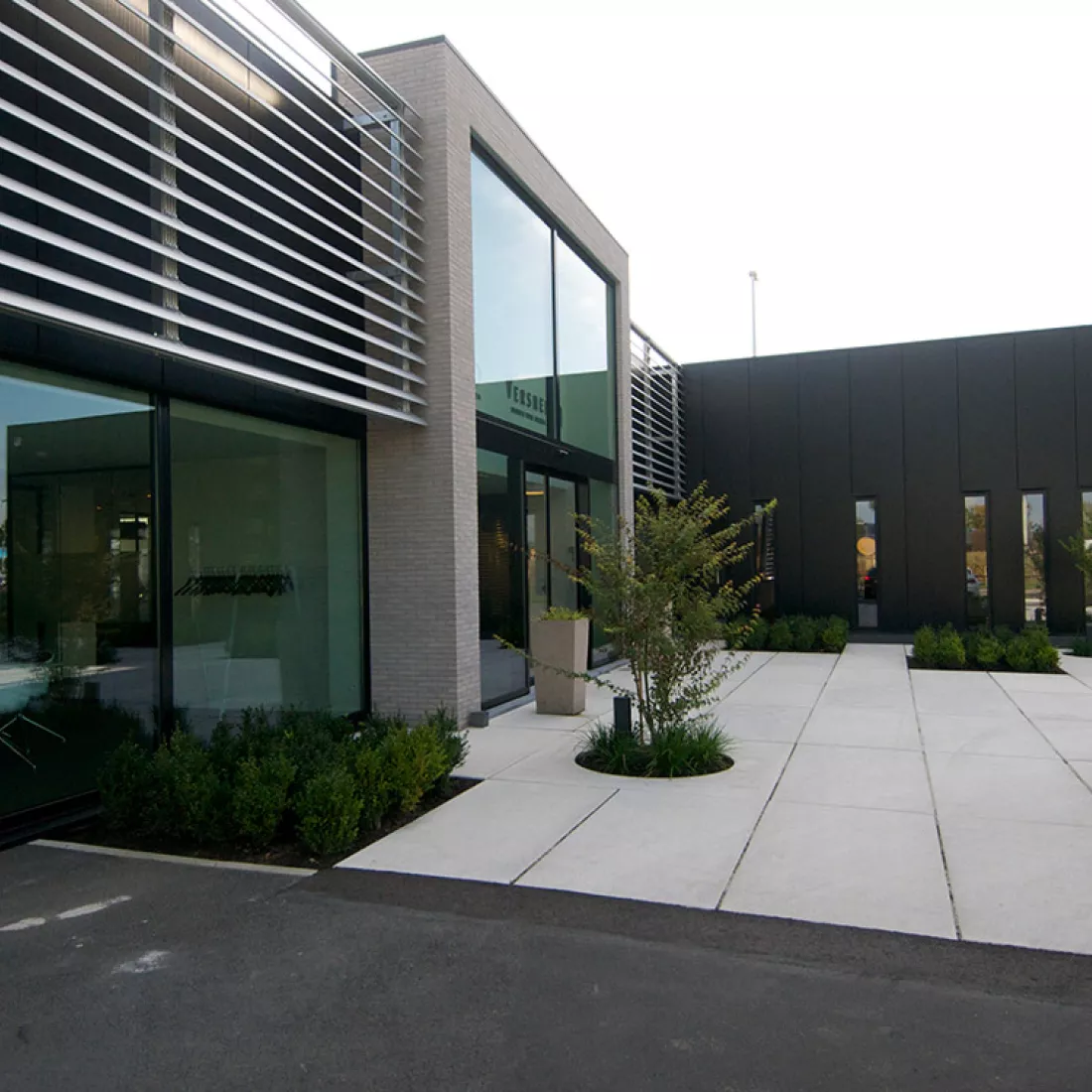 Zijaanzicht ingang showroom Vimmo. De ingang bestaat uit grote ramen met er rond een grijze steen. Naast de omranding van de grijze steen bestaan de muren volledig uit ramen.