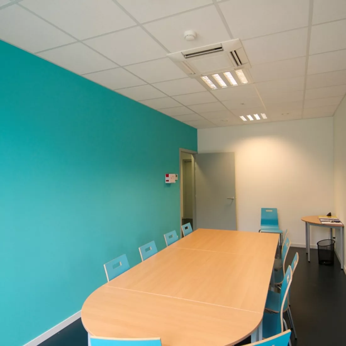 Vergader- of lunchruimte van het kinderdagverblijf. In het midden staat een grote tafel met er rond blauwe stoelen. De linker muur heeft deze zelfde blauwe kleur. Achteraan is een grijze deur.