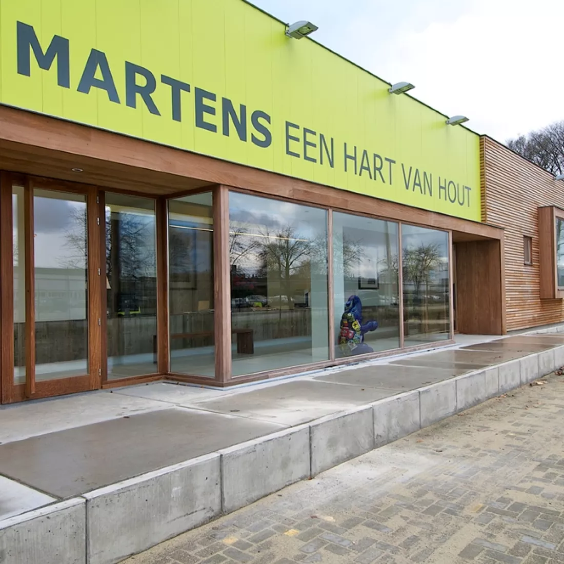 Inkom toonzaal Martens Hout. Boven de deur hangt het logo. De inkom bestaat volledig uit ramen.