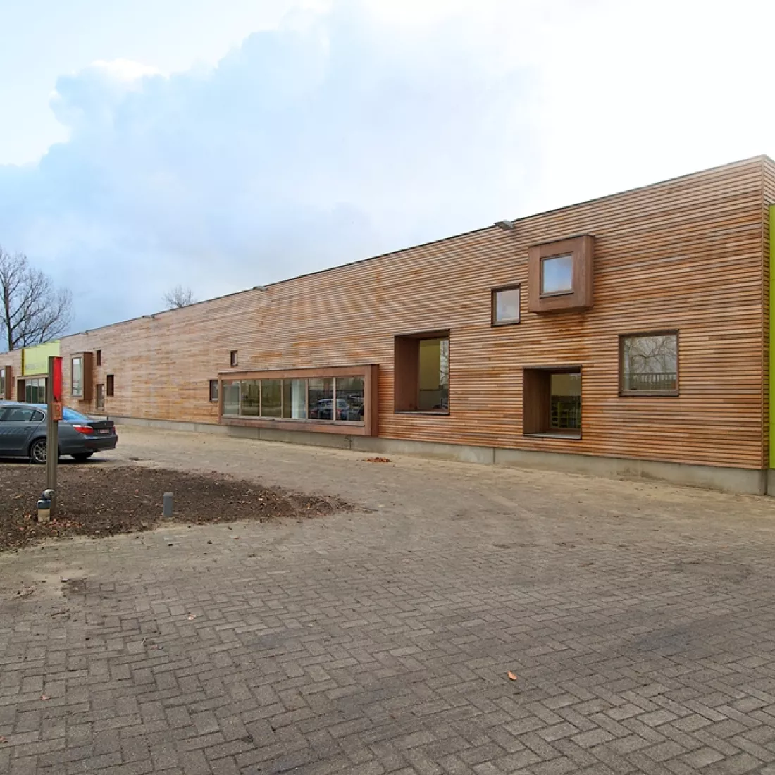 Zijaanzicht toonzaal Martens Hout. Het gebouw bevat veel kleine ramen. Links op de afbeelding en dus voor het gebouw bevinden zich parkeerplaatsen.