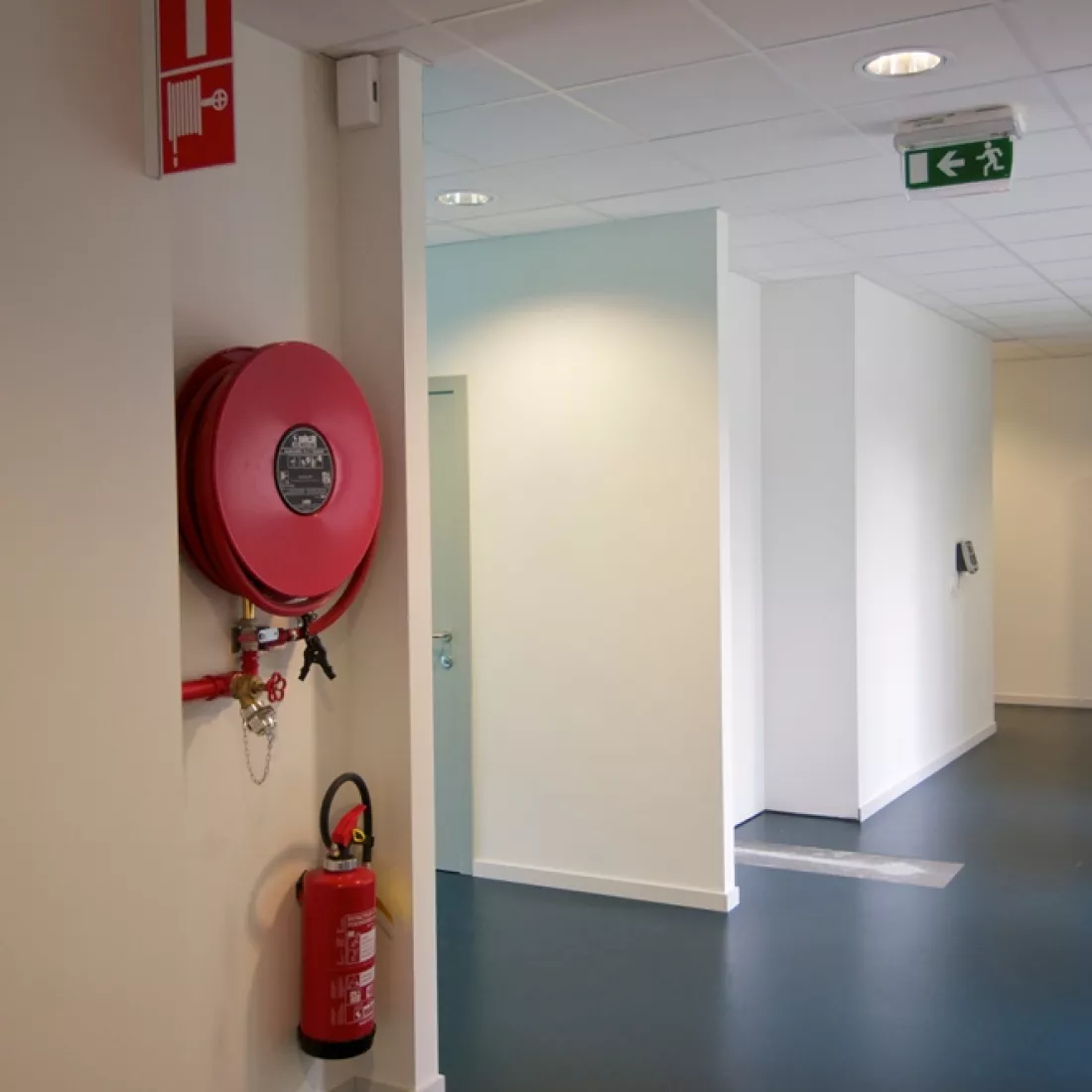 Binnenkant kinderdagverblijf. Links hangt een rode brandblusser en brandslang. Dan is er een gang naar links. De muren zijn wit en de vloer is zwart.