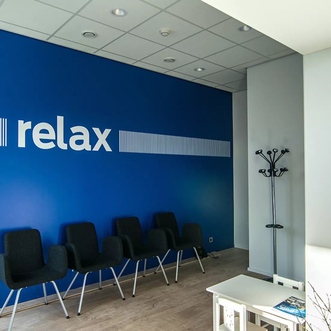 Wachtruimte garage Geudens. De linker muur is donkerblauw en bevat witte strepen en letters die het woord 'relax' vormen. Tegen de muur staan zwarte stoelen. De andere muren zijn wit. Er staat ook nog een kapstok in de ruimte en een kleine tafel met twee stoelen voor kinderen.