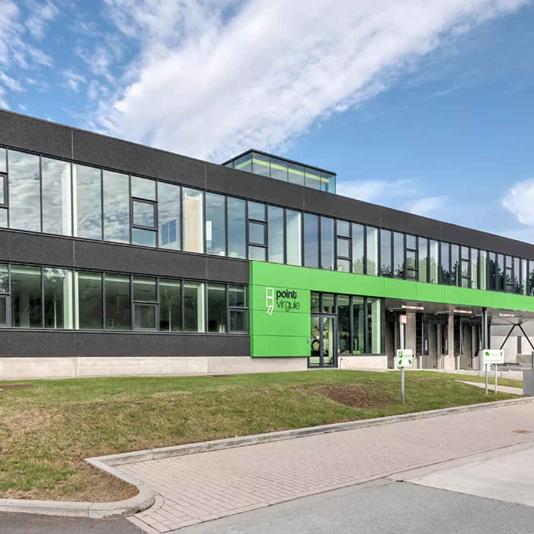 Zijaanzicht stapelmagazijn en toonzaal Point Virgule. Het gebouw heeft een zwarte kleur met groene accenten. Er zijn veel stalen ramen. Voor het gebouw is een grasveld en parking voorzien.