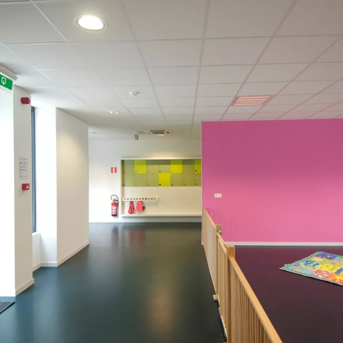 Binnenkant kinderdagverblijf. Het gebouw is opgebouwd met staal. Er is een roze muur en witte muren. Links bevinden zich twee ramen.