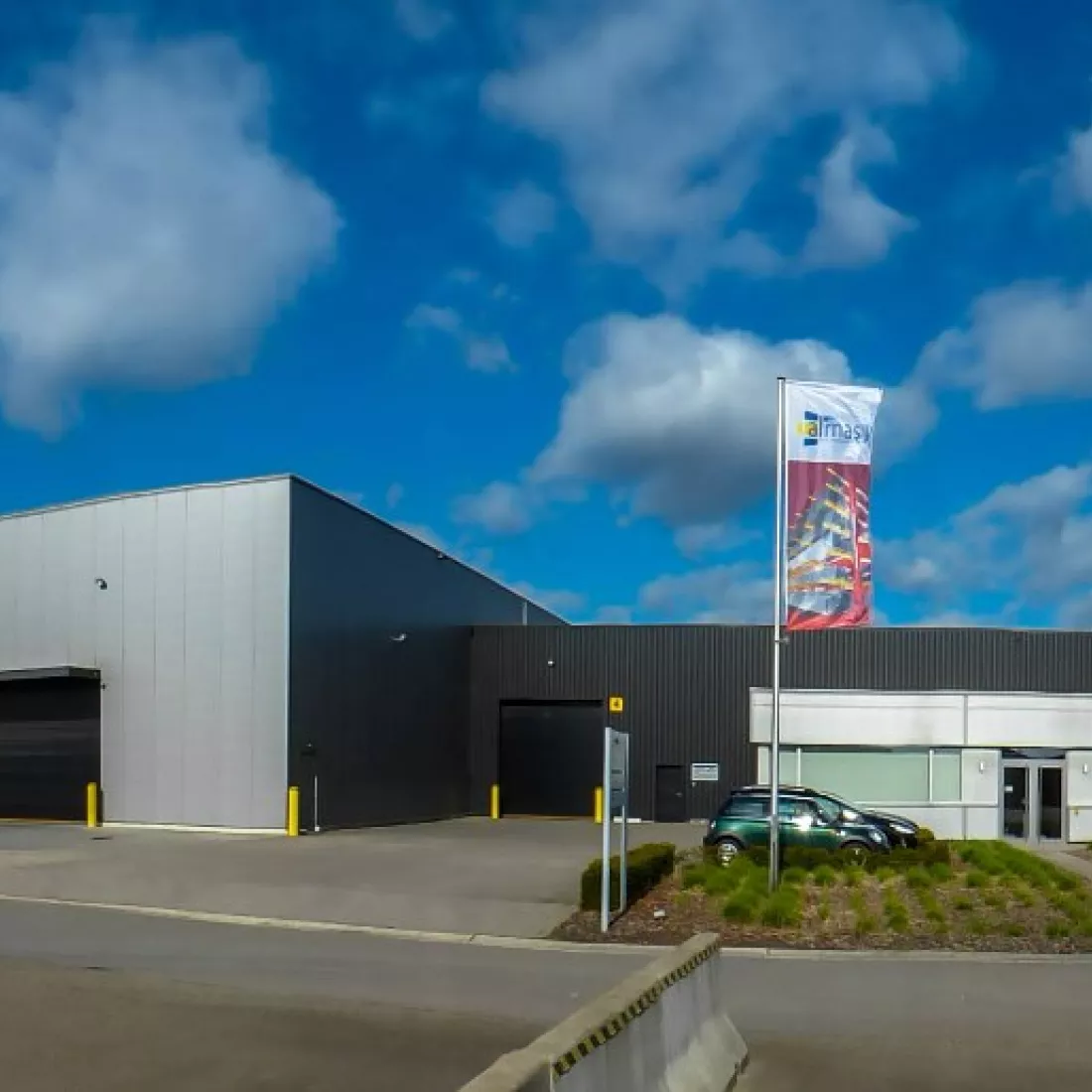 Vooraanzicht industriehal Almasy. De hal is grijs met zwart. Ervoor is parkeerruimte voorzien. Ook is er een vlag van Almasy voor de hal.