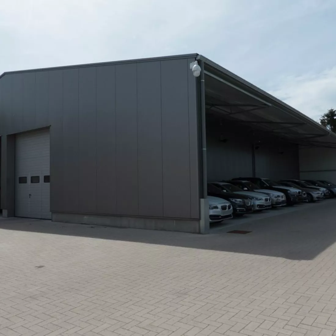 Zijaanzicht bandenloods garage Meeusen. Het gebouw is zwart en bevat een grijze poort. Rechts staan auto's geparkeerd onder de luifel.