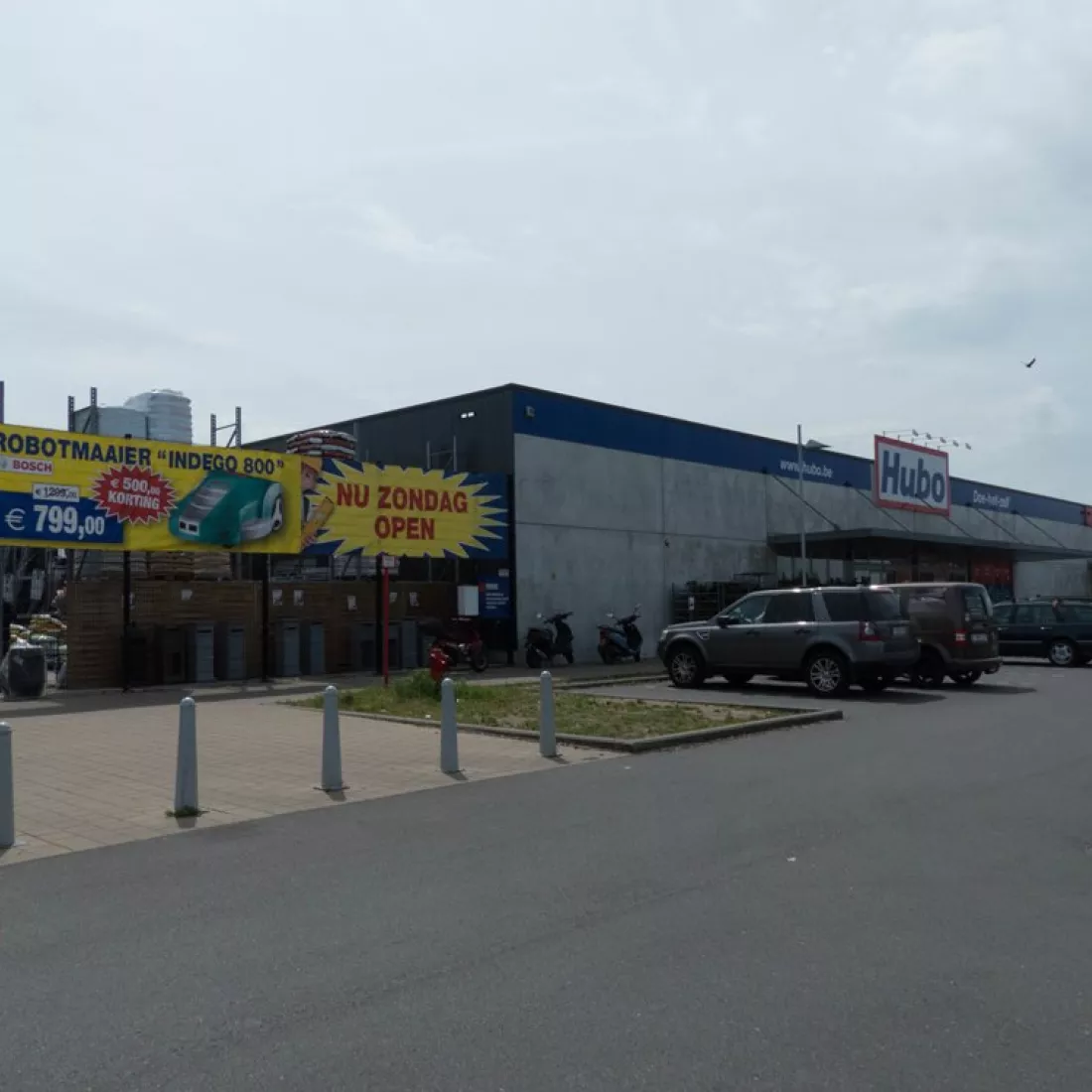 KMO Units Picolo - Van Ende: Hubo. Tijdens de opbouw zijn er verschillende materialen gebruikt zoals beton. Boven de inkom hangt een groot logo van Hubo. Voor de winkel zijn parkeerplaatsen voorzien. Links bevindt zich een hek.