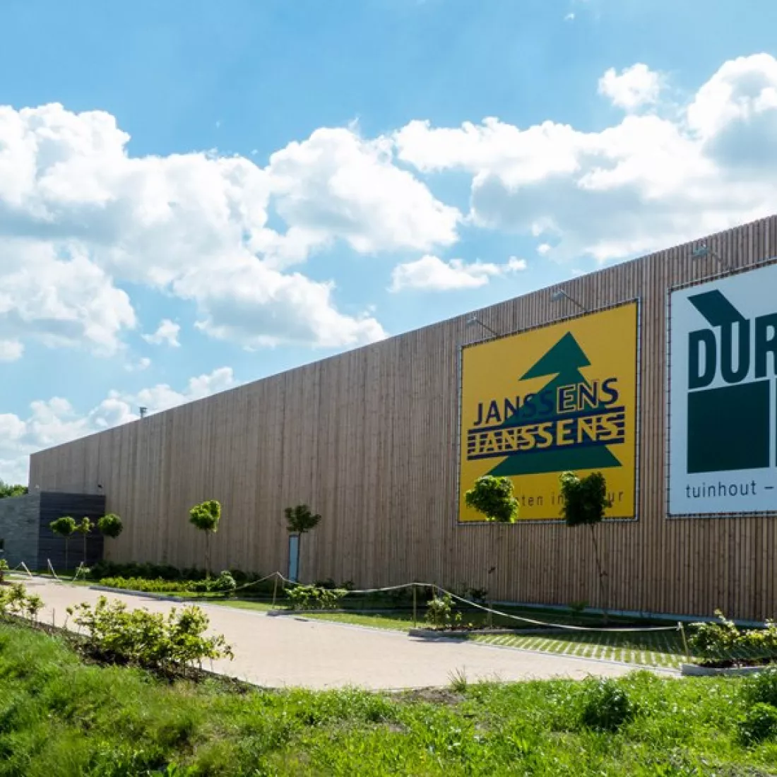 Vooraanzicht Houthandel Janssens. Het gebouw bestaat uit een combinatie van materialen, zoals hout. Rechts hangt het logo. Voor het gebouw zijn parkeerplaatsen voorzien met groen rond.