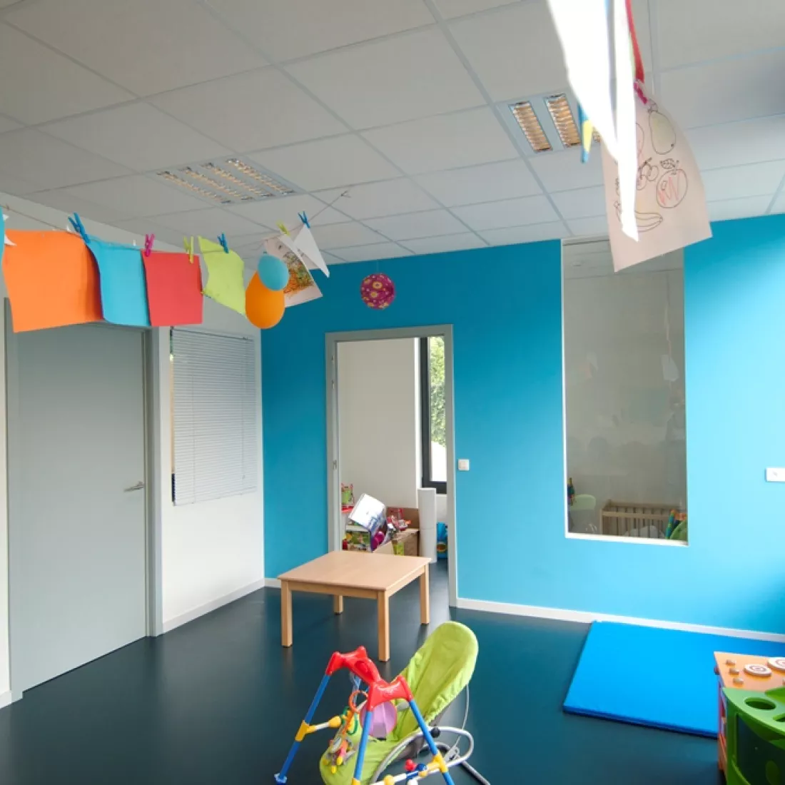 Speelruimte kinderdagverblijf. De linker muur is wit en heeft een grijze deur. De muur ernaast heeft een blauwe kleur en heeft ook een grijze deur en een raam om naar de volgende ruimte te kijken. Er hangen papieren aan een draad. Deze draad is bevestigd aan het plafond. In de ruimte staan kinderspullen.