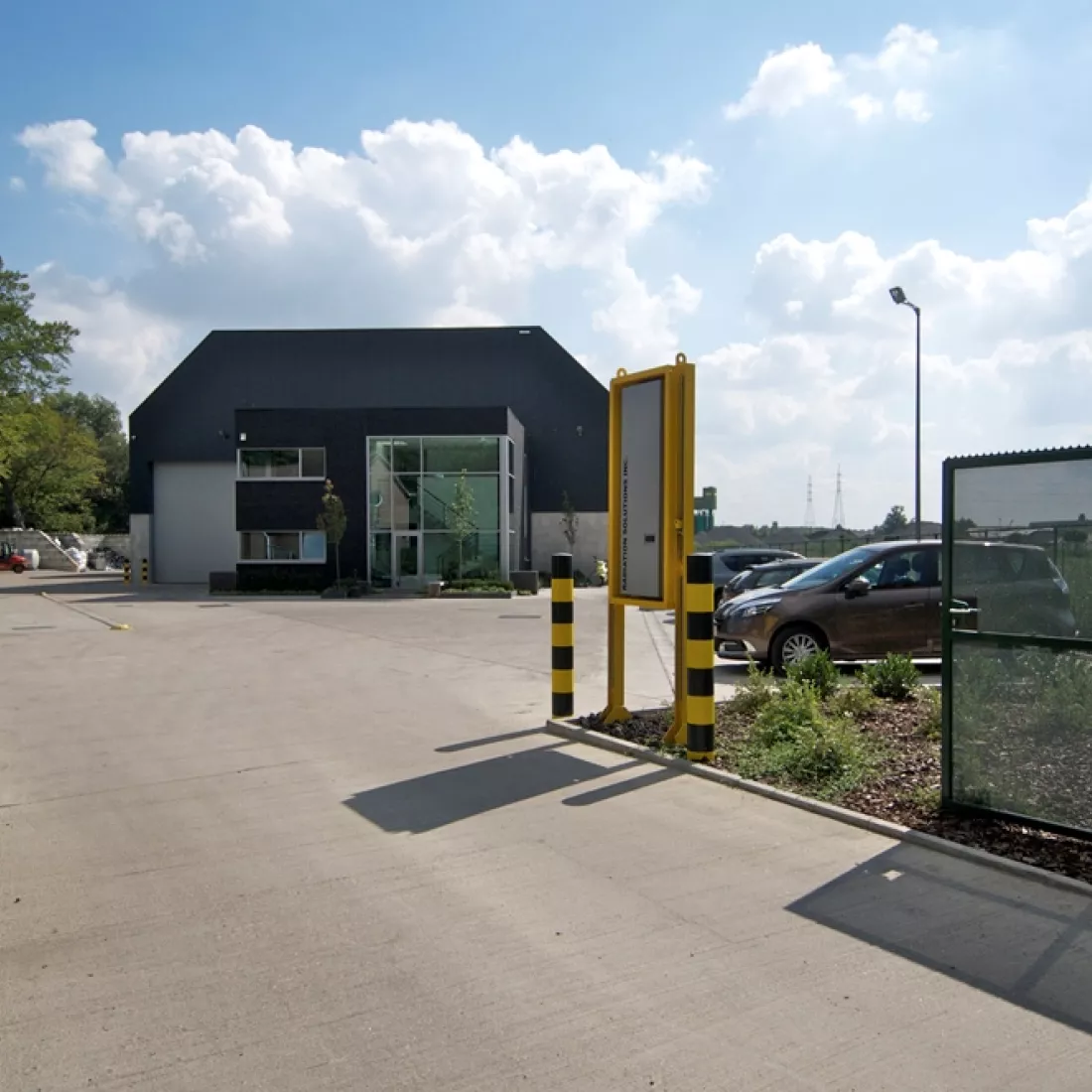 Vooraanzicht kantoorgebouw en opslagplaats GRSE. Het kantoorgebouw bevindt zich voor de opslagplaats. Het kantoorgebouw is zwart en de opslagplaats is onderaan grijs. Bovenaan bestaat de opslagplaats uit donkerblauwe platen. Het linkerdeel van de opslagplaats bevat een grote grijze poort.