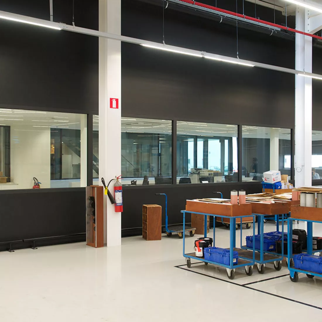 Binnenkant productiehal - Reynesco. Rechts staan werkmaterialen in afgebakende vakken. De vakken zijn afgebakend met zwarte lijnen. Links bevinden zich bureaus. De bureaus zijn achter een muur met ramen in. De muur is zwart.