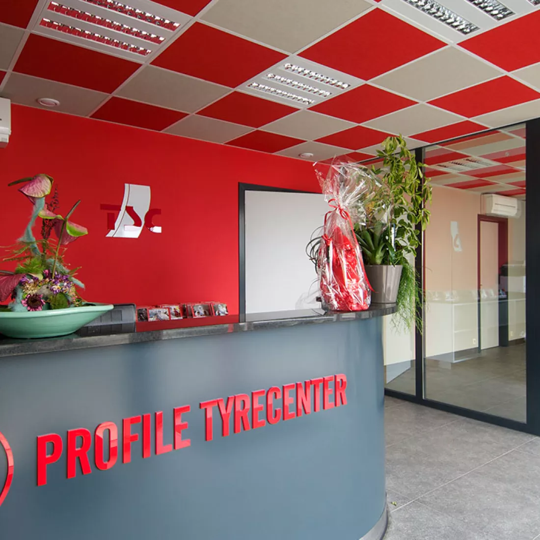 Binnenkant inkom - TSC. Er staat een grijze balie waar met rode letters "Profile typecenter" opstaat. De muur achter de balie is rood en tegen deze muur hangt het logo van TSC. Rechts bevindt zich een witte deur. Rechts bevindt zich ook een glazen wand. Het plafond bestaat uit rode en witte vierkanten.
