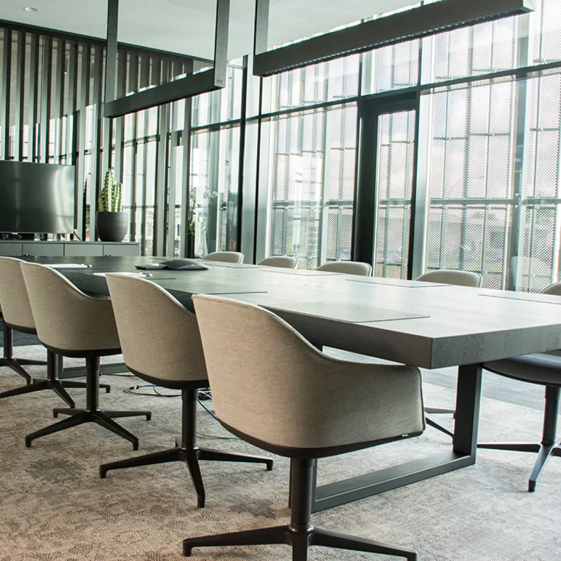 Binnenkant vergaderruimte Power Solutions. In het midden staat een lange rechthoekige tafel met acht grijze stoelen. Onder de tafel ligt een tapijt. Links daarvan staat een TV en een plant. Achter de tafel bestaat de muur uit grote ramen.