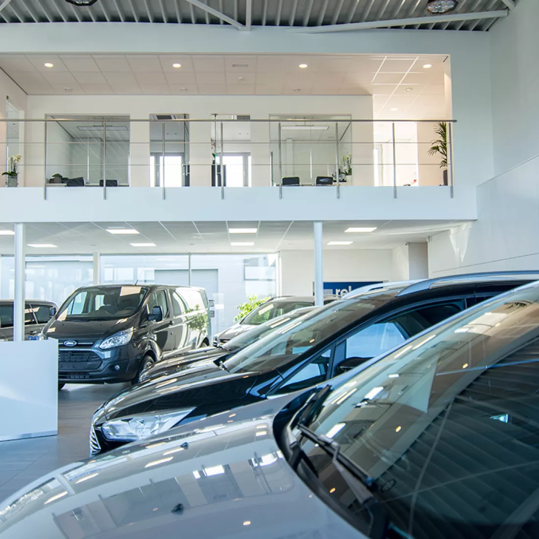 Binnenkant showroom en kantoor garage Geudens. Op de gelijkvloers staan de auto's in de showroom. Achteraan is een railing voor de eerste verdieping. Daar bevinden zich de kantoren.