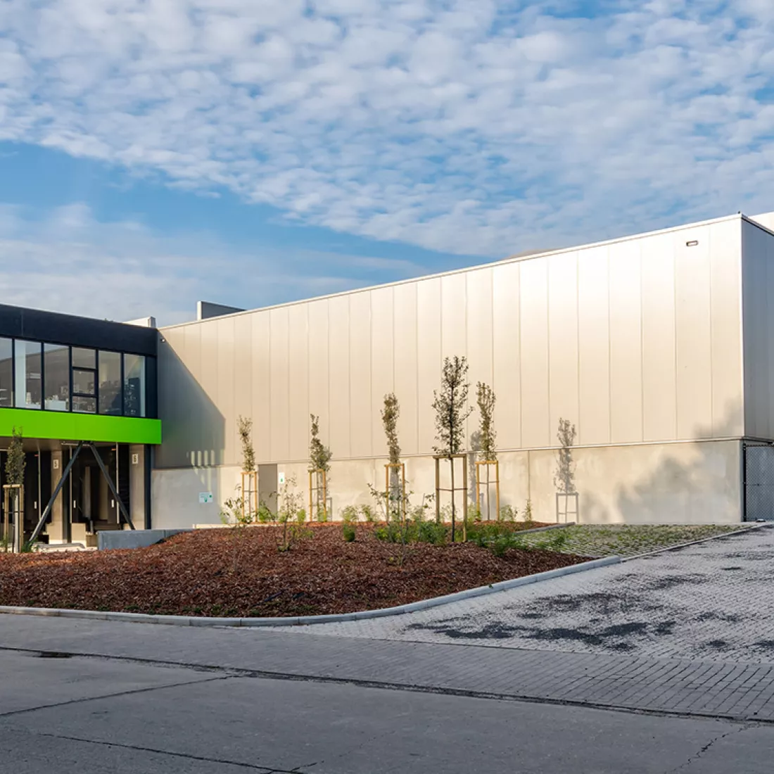 Zijaanzicht stapelmagazijn en toonzaal Point Virgule. Het gebouw is gemaakt uit staal. Het heeft een grijze, zwarte en groene kleur. Ervoor staan enkele bomen.