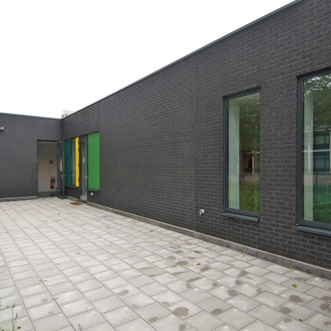 Zijaanzicht buitenkant kinderdagverblijf. Het gebouw bestaat uit zwarte steen en ramen met groene en gele details. Er zijn ook deuren aanwezig.