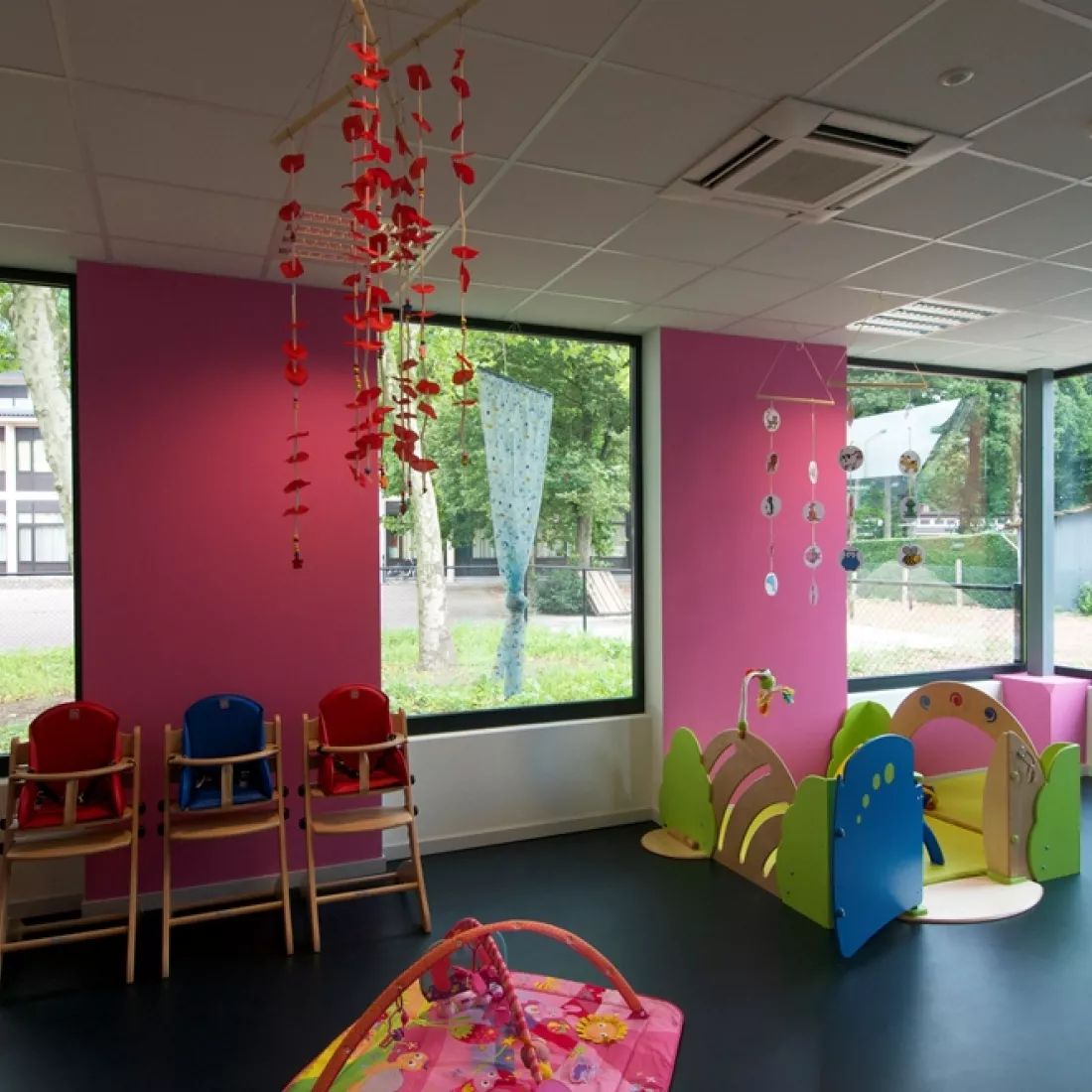 Speelruimte kinderdagverblijf. De muren zijn roos en hebben grote ramen om naar buiten te kijken. Er staan kinderstoelen en - speelgoed.