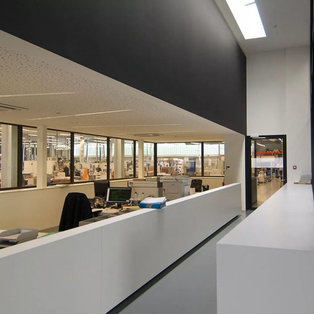Binnenkant kantoor productiehal - Reynesco. Links staan er bureaus. Rond de bureaus is een muur met ramen in. De ramen hebben zicht op de productiehal. Rechts naast de bureaus staat een halve muur, dan is er een gang. Op het einde van de gang is een deur.