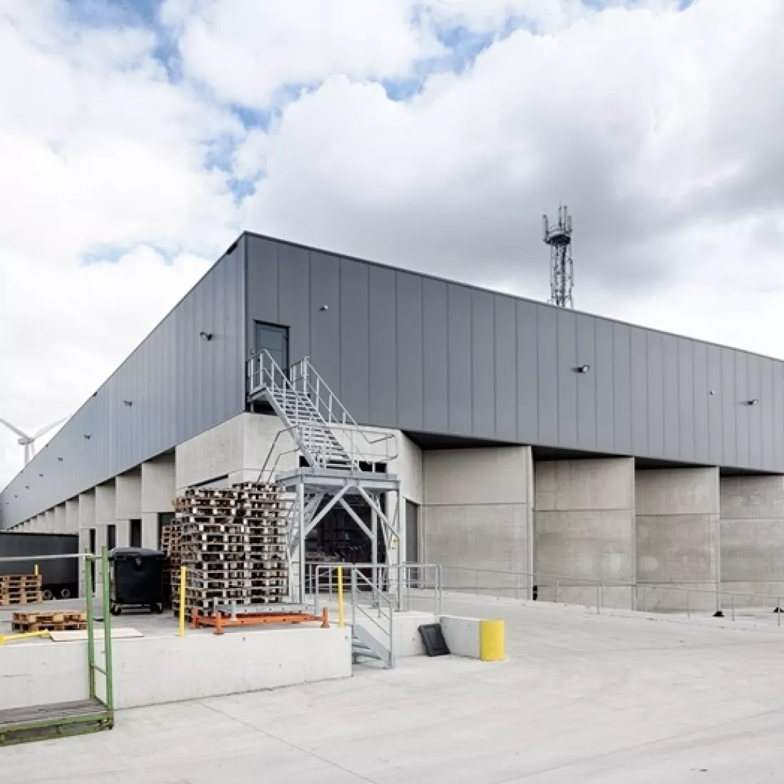 Zijaanzicht transitmagazijn Transuniverse. De onderkant van het gebouw bestaat uit beton en de bovenkant is donkergrijs. Er gaat langs buiten een trap naar een deur.