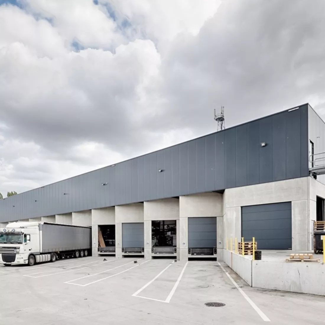 Dock magazijn Transuniverse voor het laden en lossen van vrachtwagens. De poorten zijn donkergrijs net zoals de bovenkant van het magazijn. De onderkant bestaat uit beton.