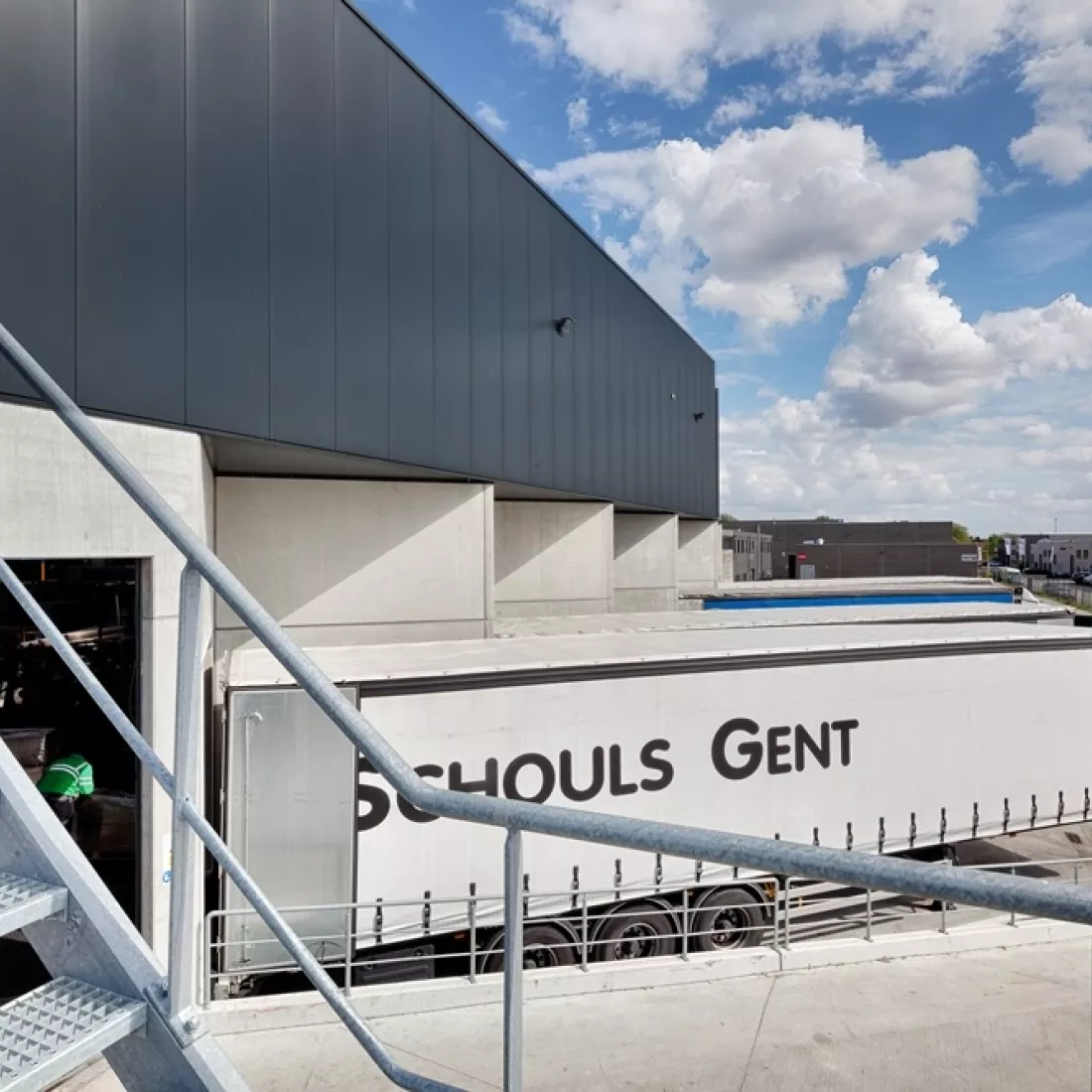 Zijaanzicht dock voor het laden en lossen van vrachtwagens. De bovenkant van het gebouw is donkergrijs en de onderkant bestaat uit beton. Er staan enkele vrachtwagens te laden.