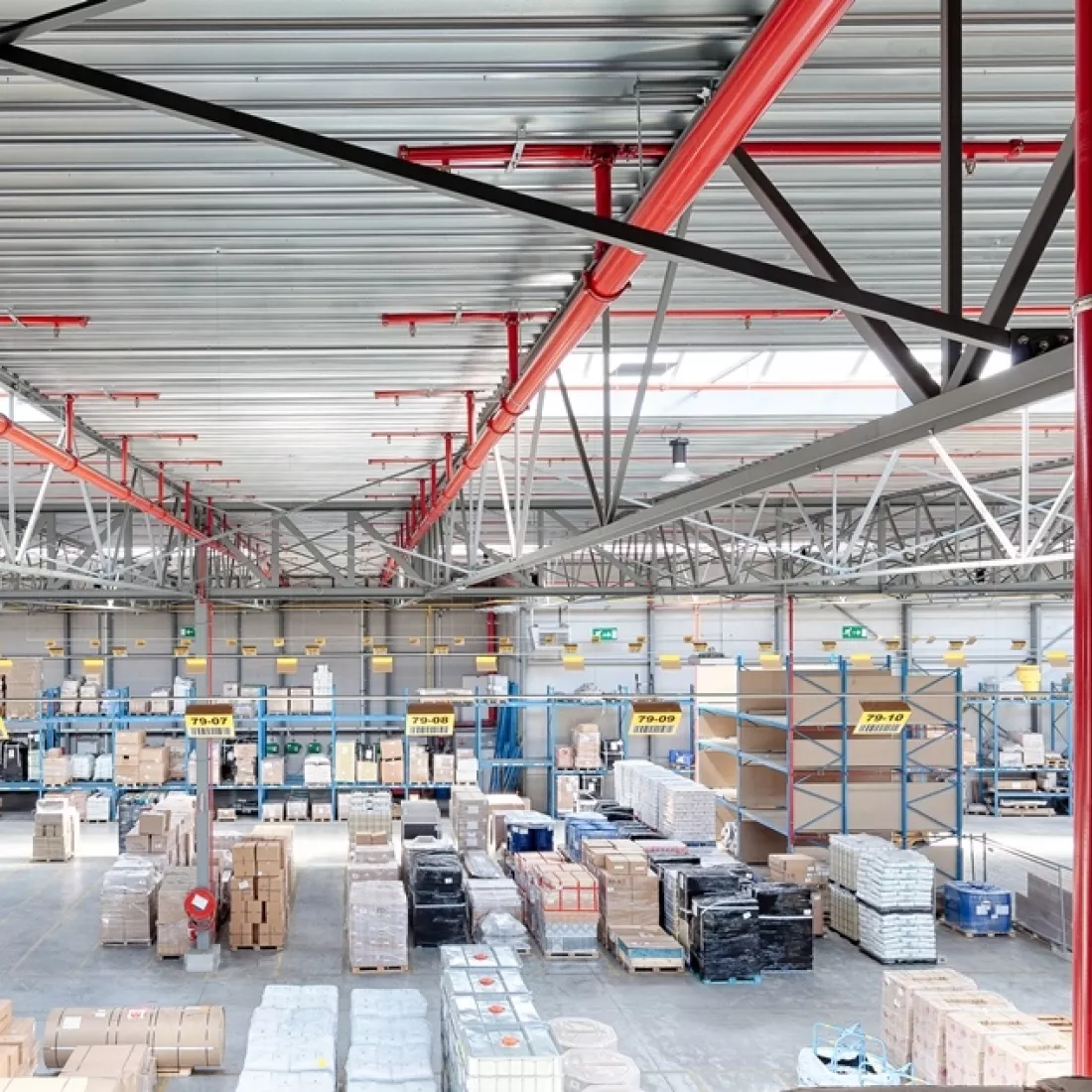 Binnenkant transitmagazijn Transuniverse. Bij de opbouw is gebruik gemaakt van staal. Nu staan er verschillende producten opgeslagen zowel in rekken als los. Aan het dak hangen rode buizen.