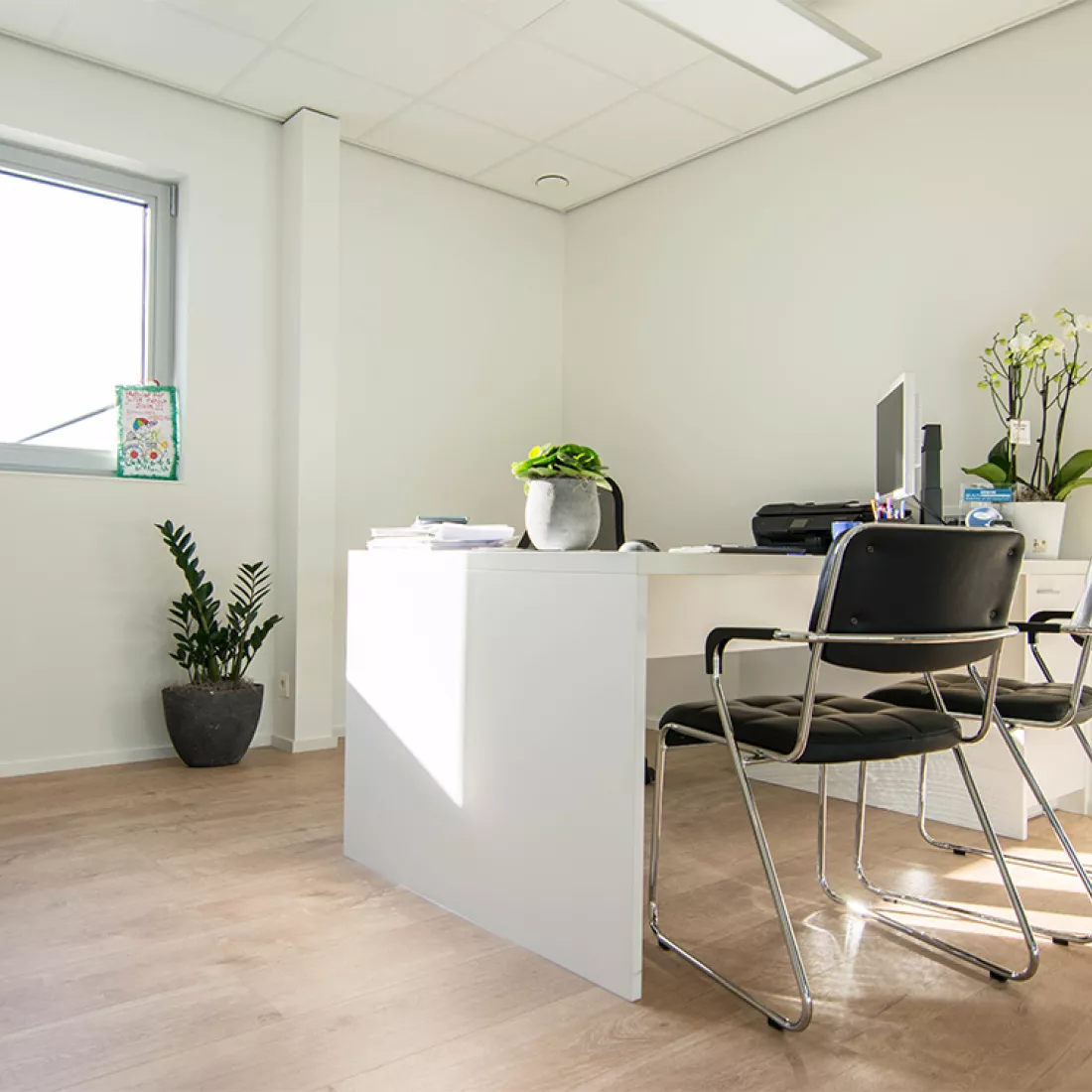 Close-up kantoor garage Geudens. De witte bureau staat tegen de rechtermuur. Links van de bureau staat een bureaustoel met computerscherm. Rechts van de bureau staan twee zwarte stoelen. De muur achter de bureau heeft een raam.
