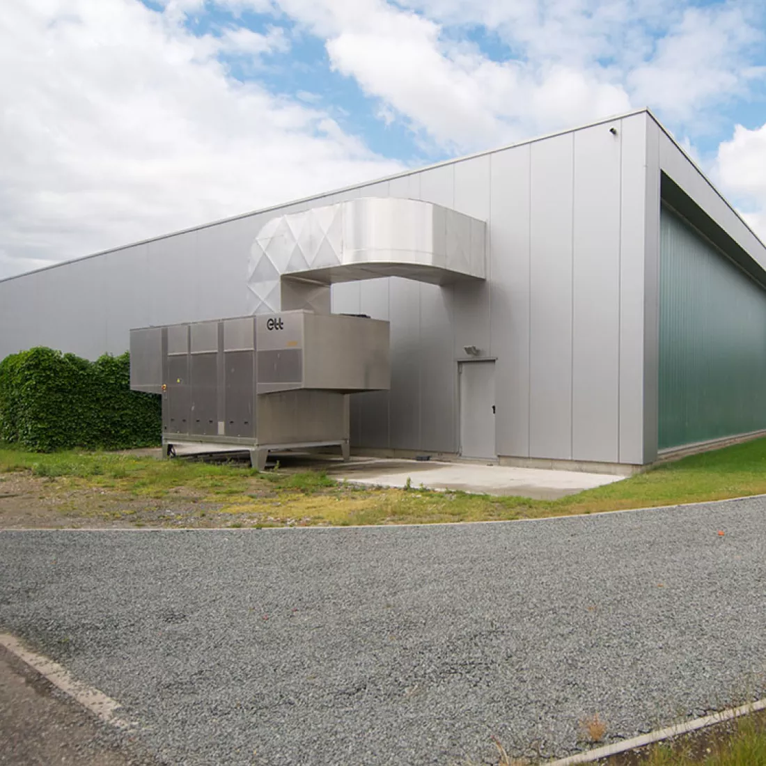 Achterkant productiehal - Reynesco. Het gebouw bestaat uit grijze platen. De achterkant heeft ook een grijze deur. De rechtermuur bevat een groot raam. Het raam is niet volledig doorzichtig.