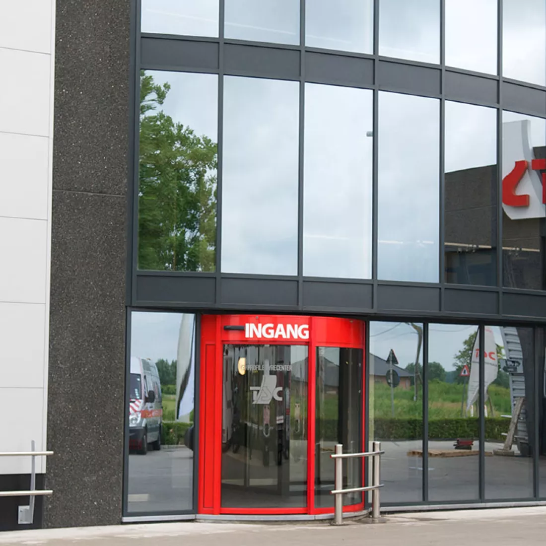 Ingang - TSC. De ingang bestaat uit een rode draaideur met in het wit het woord "ingang" erop. Dit deel van het gebouw bestaat uit grote ramen met spiegelglas. Het gebouw heeft drie verdiepingen.