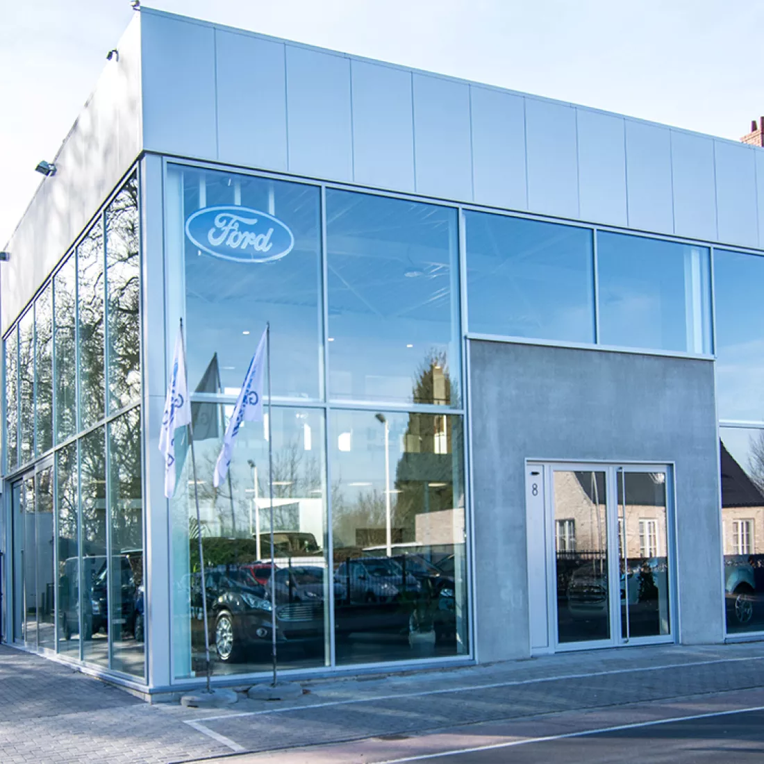 Zijaanzicht showroom garage Geudens. Het gebouw is grijs en bevat veel ramen om de auto's in de showroom te zien staan. In de linkerhoek binnen hangt het logo van Ford.