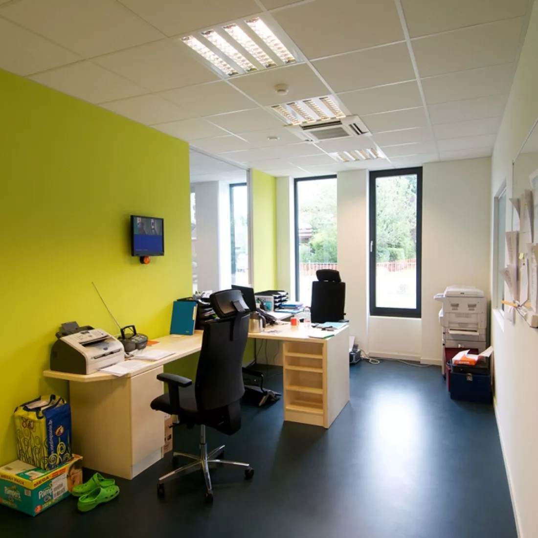Kantoorruimte in kinderdagverblijf. De bureau staat tegen de linker muur, die heeft een groene kleur. Rechts van het bureau is een buitenmuur met ramen. Naast het raam staat een printer.