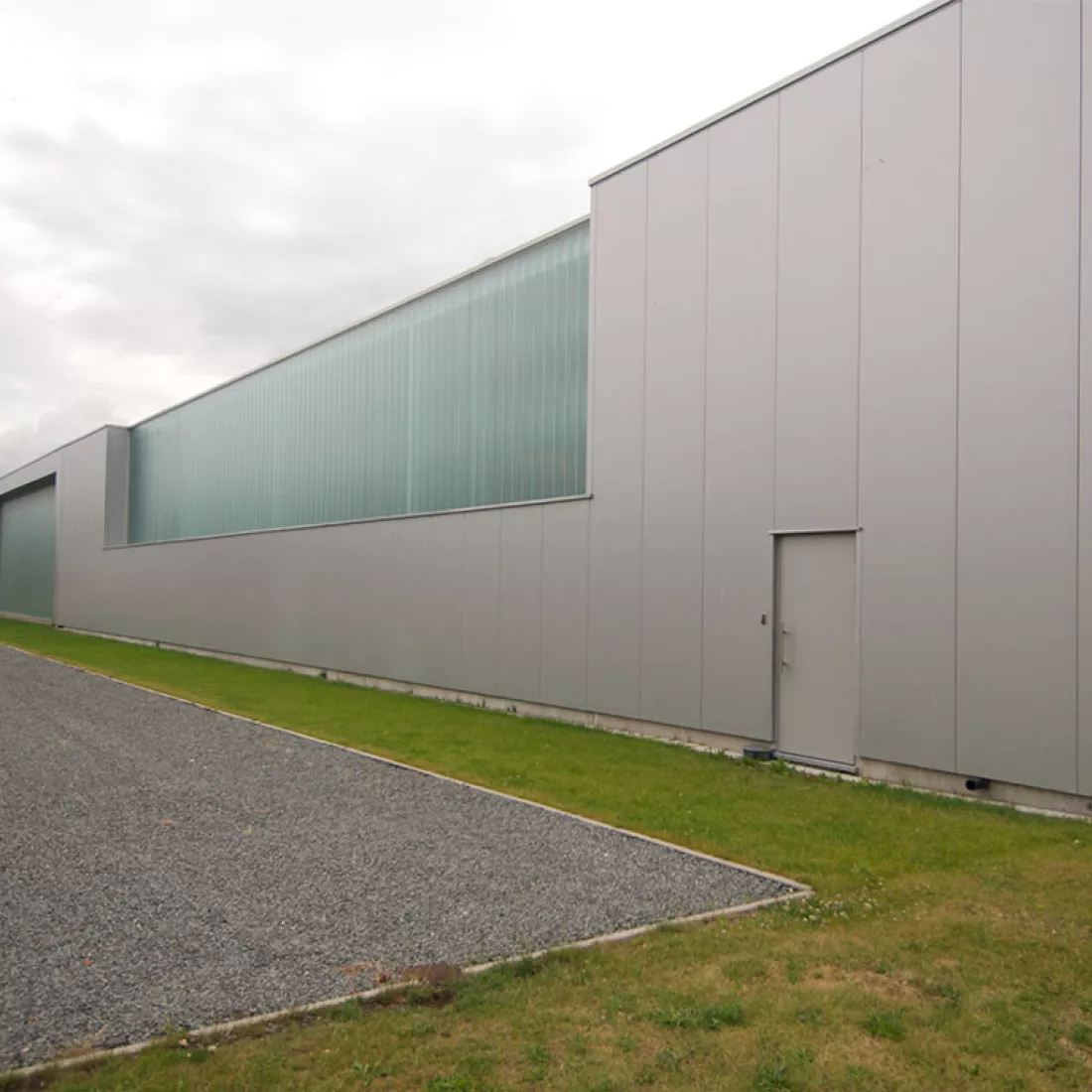 Zijaanzicht productiehal - Reynesco. Het gebouw bestaat uit grijze platen en bevat een grijze deur. Ook bevat het gebouw grote ramen die niet volledig doorzichtig zijn. Rond het gebouw bevindt zich gras en daarna een kiezelpad.