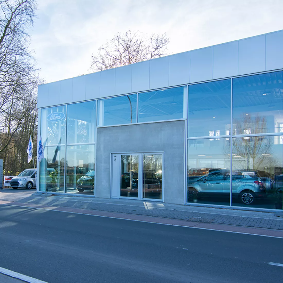 Vooraanzicht showroom garage Geudens. Het gebouw is grijs en bevat veel ramen om de auto's in de showroom te zien staan. De afbeelding is genomen van de overkant van de straat.